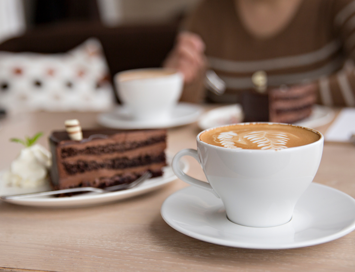 Kaffeetasse mit Latte-Art und Schokoladenstückchen auf einem Tisch, im Hintergrund eine Person mit Streifenpullover.