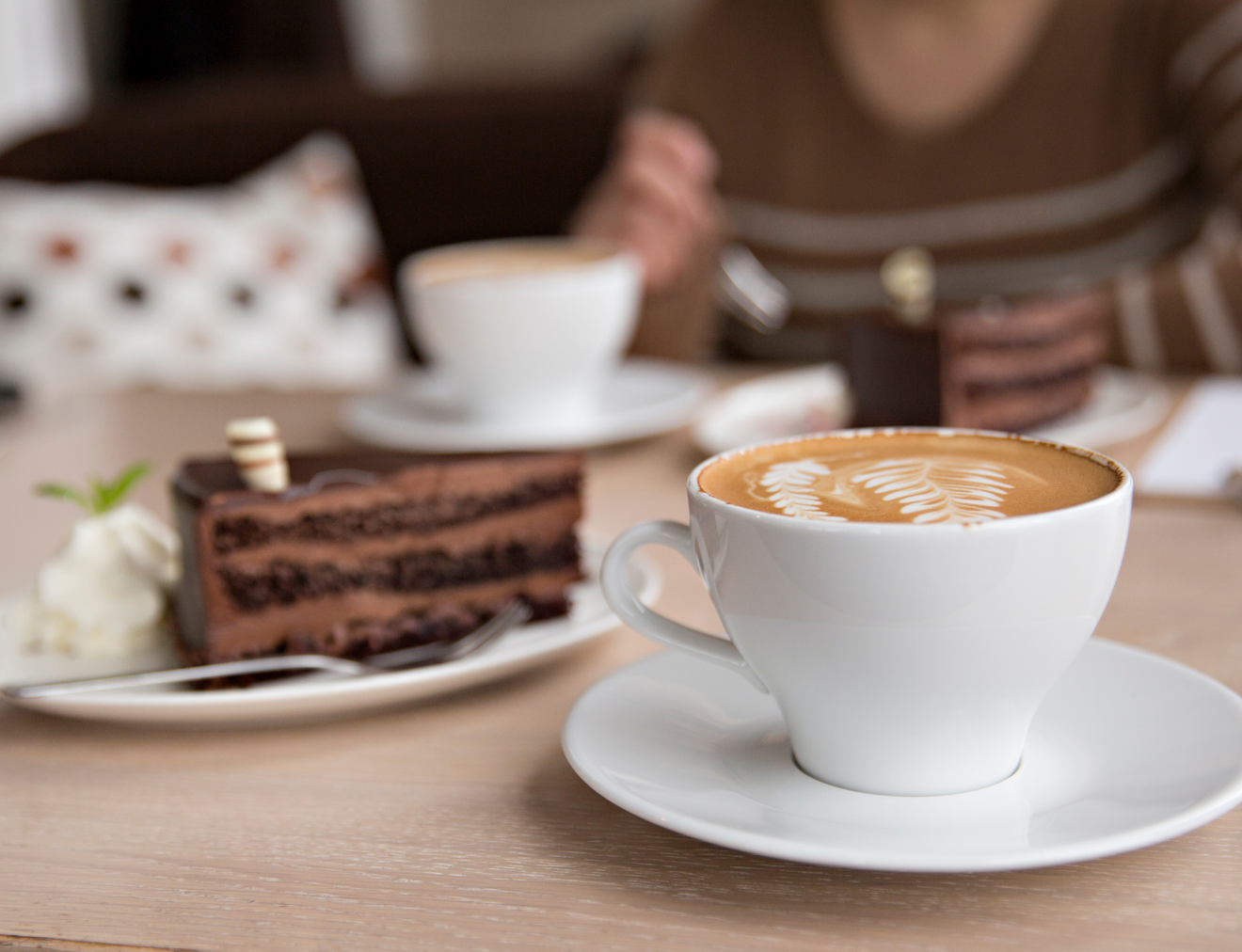 Kaffeetasse mit Latte-Art und Schokoladenstückchen auf einem Tisch, im Hintergrund eine Person mit Streifenpullover.
